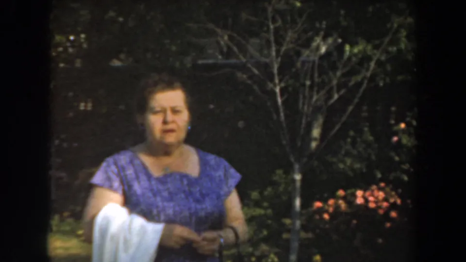 The woman faces the camera in a 1961 Los Angeles garden scene, with flowering bushes and a small tree behind her.