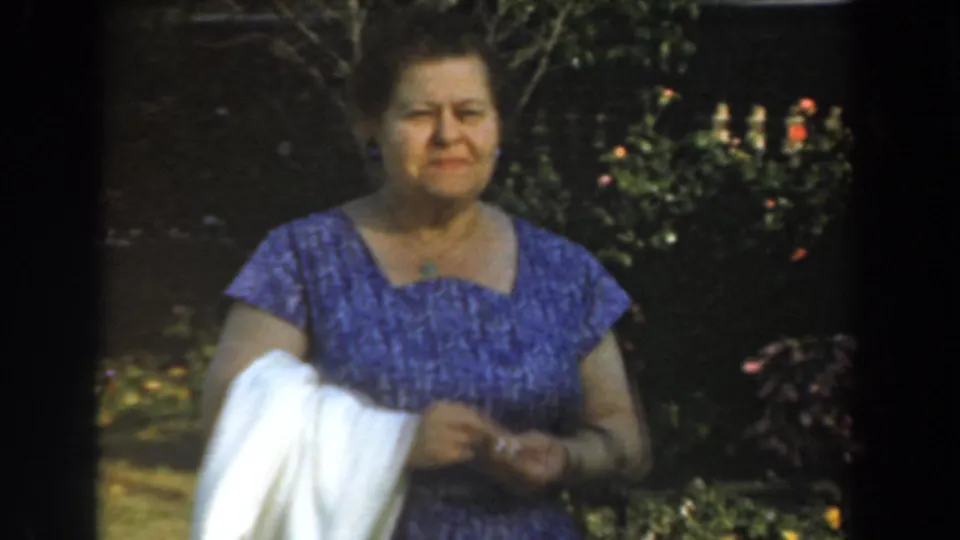 Adult woman in a purple patterned dress stands outdoors in Los Angeles 1961 home-movie footage, holding a white cloth near garden foliage.