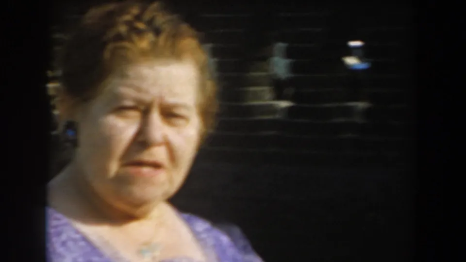 Close handheld frame of an adult woman in a purple patterned dress against a dark fence or wall in 1961 Los Angeles home-movie footage.
