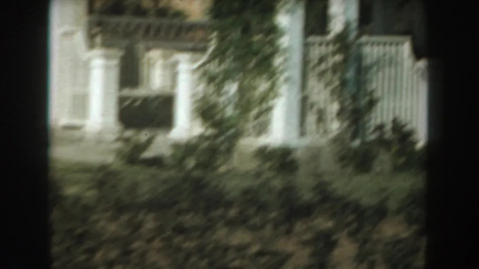 A blurry 1961 Los Angeles garden view shows a white exterior structure or fence with greenery in front.