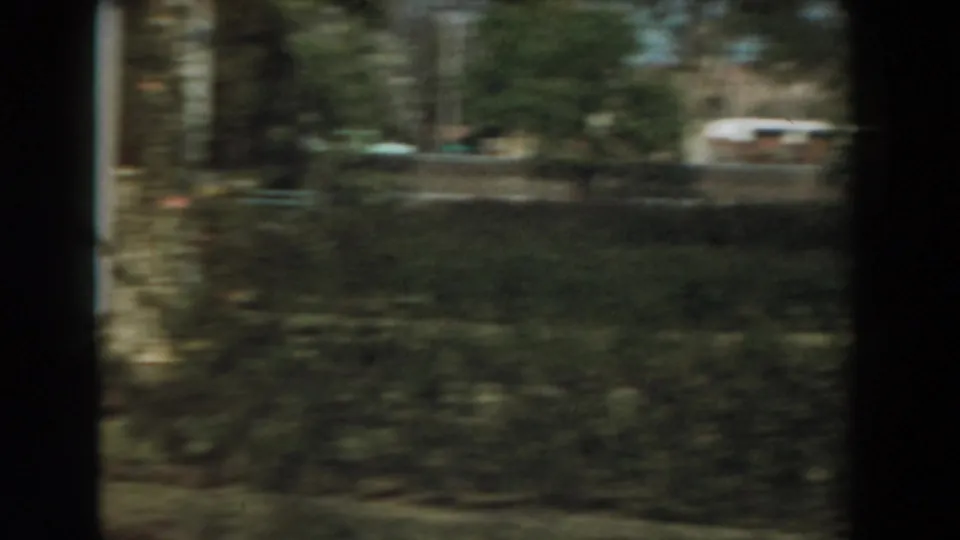 A handheld 8mm frame shows a blurred garden or yard with trees, grass, and a distant light-colored vehicle.