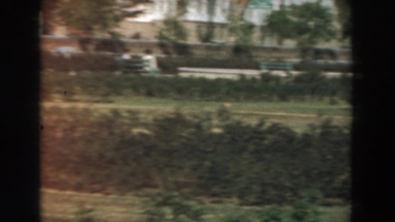 Moving view of lawns, hedges, trees, and pale buildings beyond dark film-frame edges.