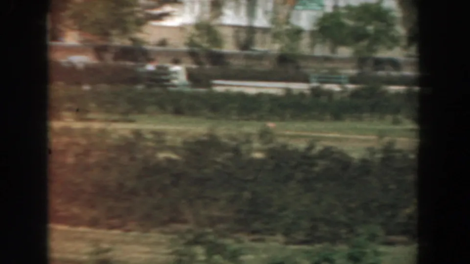 1961 Los Angeles home-movie frame showing landscaped grounds, hedges, mature trees, and distant pale buildings from a moving viewpoint.