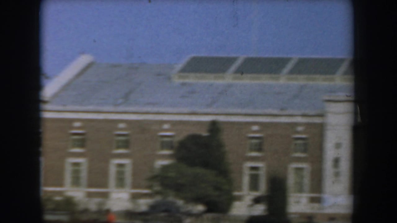 Tan building with white trim, roof vents, shrubs, and a small red figure near a path.