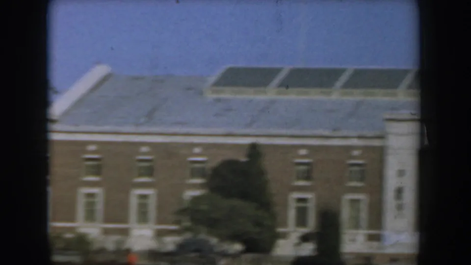 1961 Los Angeles exterior view of a tan building with white trim, roof vents, shrubs, and a distant red-coated pedestrian.