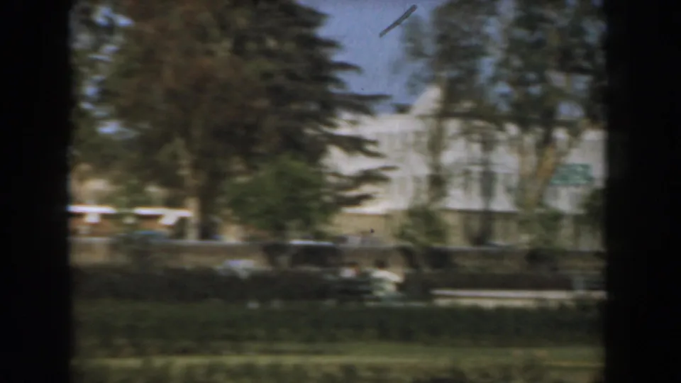 1961 Los Angeles frame showing a white building behind trees, with landscaped grounds and street-side foreground blur.
