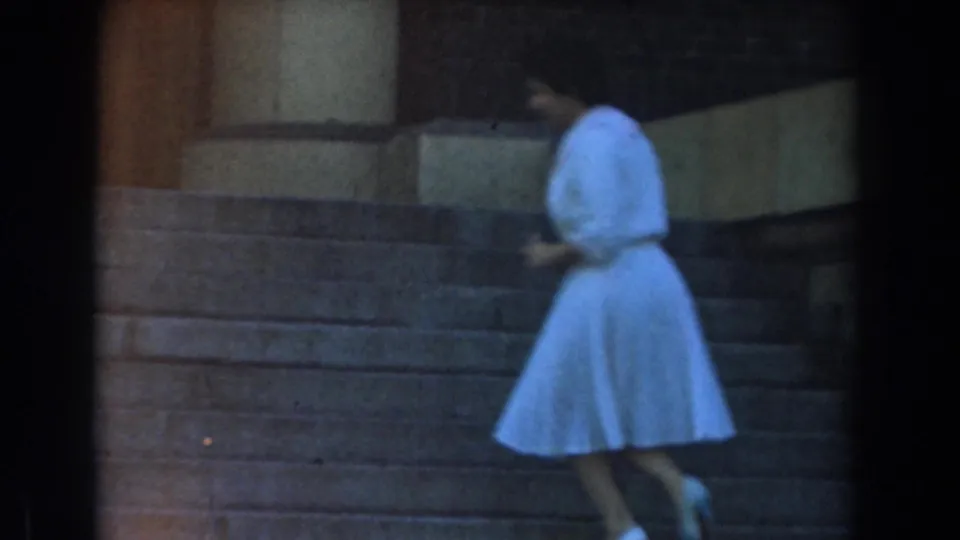 1961 Los Angeles home-movie frame of a woman in a white dress ascending stone steps and turning slightly toward the camera.