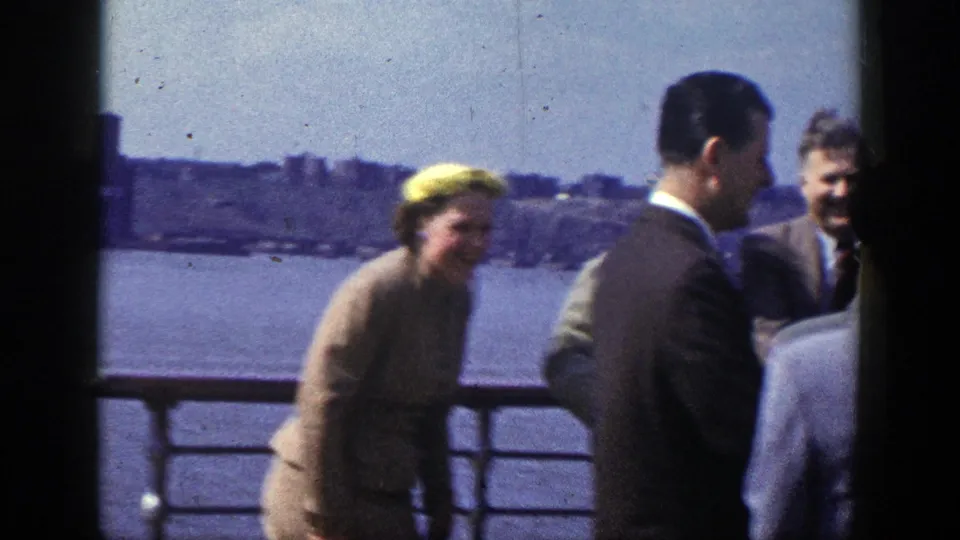 A woman in a yellow hat stands near suited men on a New York boat deck, with water and a distant shoreline behind them.