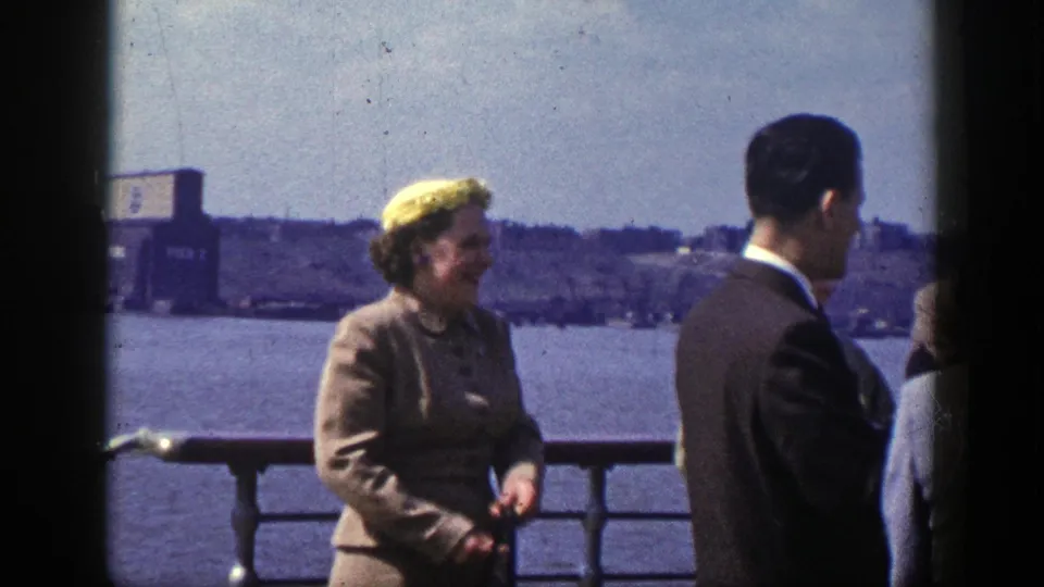 On the boat deck, a woman in a yellow hat and tailored coat stands near a suited man, with New York waterfront scenery behind them.