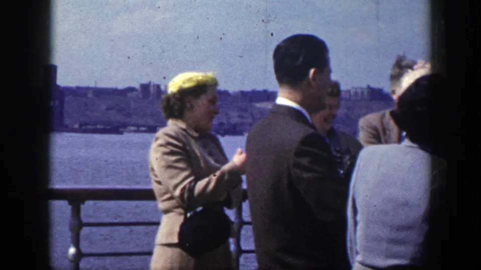Formally dressed adults on a New York boat deck converse by the railing, including a woman in a yellow hat gesturing toward the group.