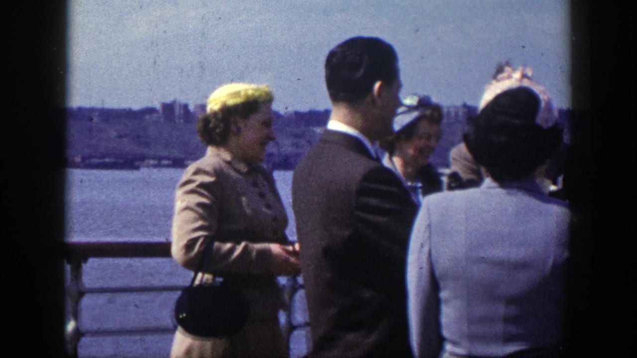 Well-dressed adults talk beside a waterfront railing with skyline across the water.