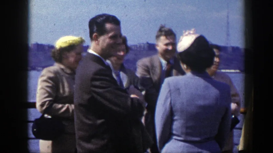 Adults in 1940s attire stand near the New York waterfront, smiling and talking with the city skyline visible across the water.