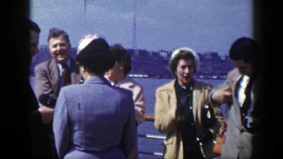 The home-movie frame shows adults in formal daywear gathered along a New York waterfront railing with blue water and skyline beyond.