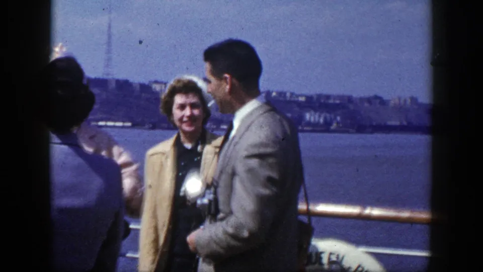A woman in a light coat and a man in a suit stand on a New York riverboat or ferry deck with water and skyline in the background.