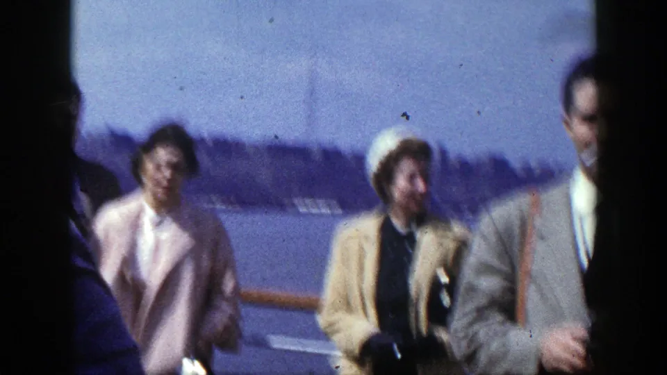 Soft-focus 8mm frame of adults in period coats on a New York boat deck, with water, railing, and shoreline visible behind them.