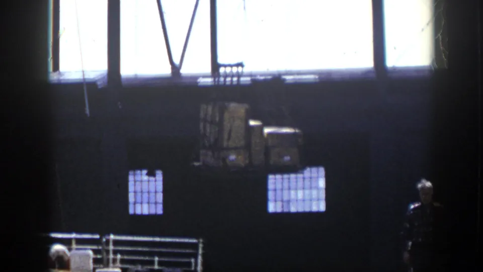 Dark waterfront industrial interior appears to show crane equipment, high windows, railings, and a person standing near the lower right of frame.