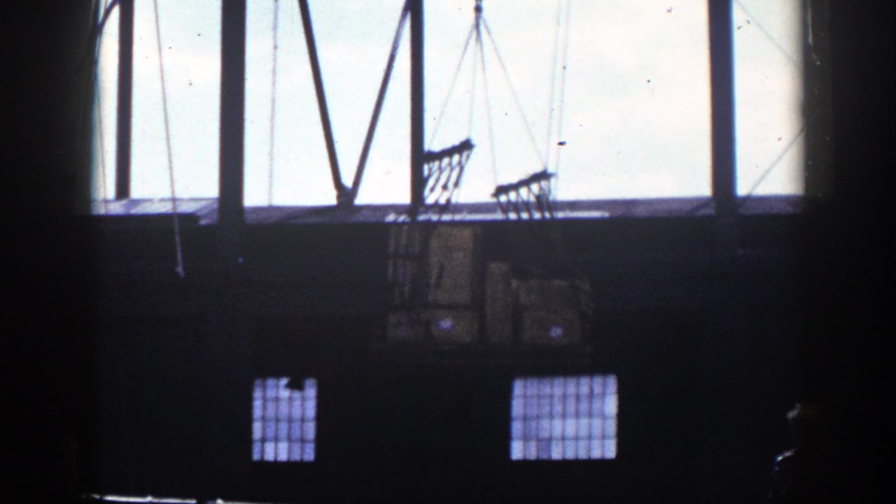Dim warehouse interior with crane cables and net-wrapped cargo boxes silhouetted against large windows.