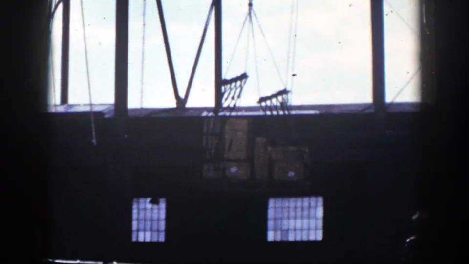 1948 New York 8mm frame showing dark warehouse space, tall windows, crane lines, and net-wrapped cargo boxes in silhouette.