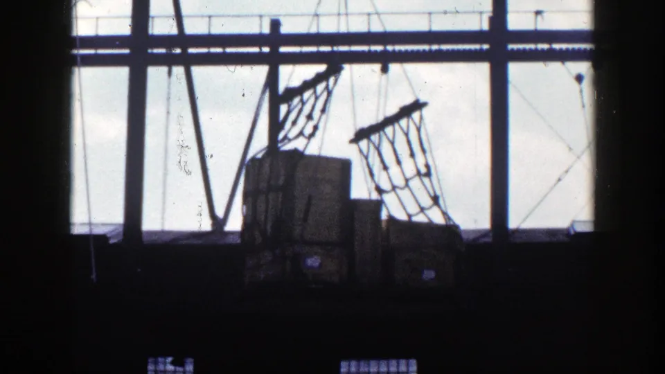 High-contrast 8mm frame of a New York warehouse, showing net-wrapped cargo boxes, cables, beams, and backlit windows.