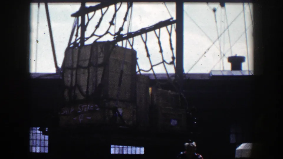 Suspended wooden crates and rope netting are seen from a dim interior at a New York dock, with bright sky through the windows.