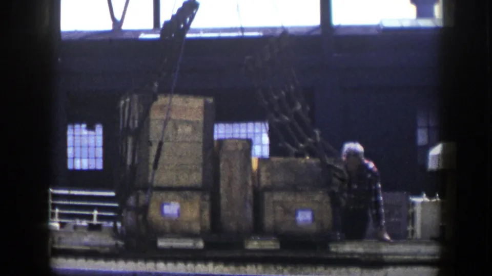 A worker in a plaid shirt appears beside wooden crates and cargo netting on a New York dock platform in the 1948 home-movie clip.