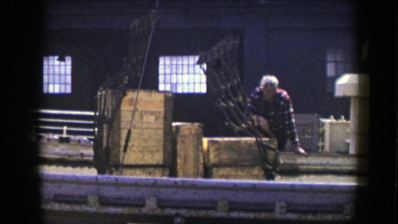 Worker in a plaid shirt bends near machinery and wooden crates inside a dim factory.