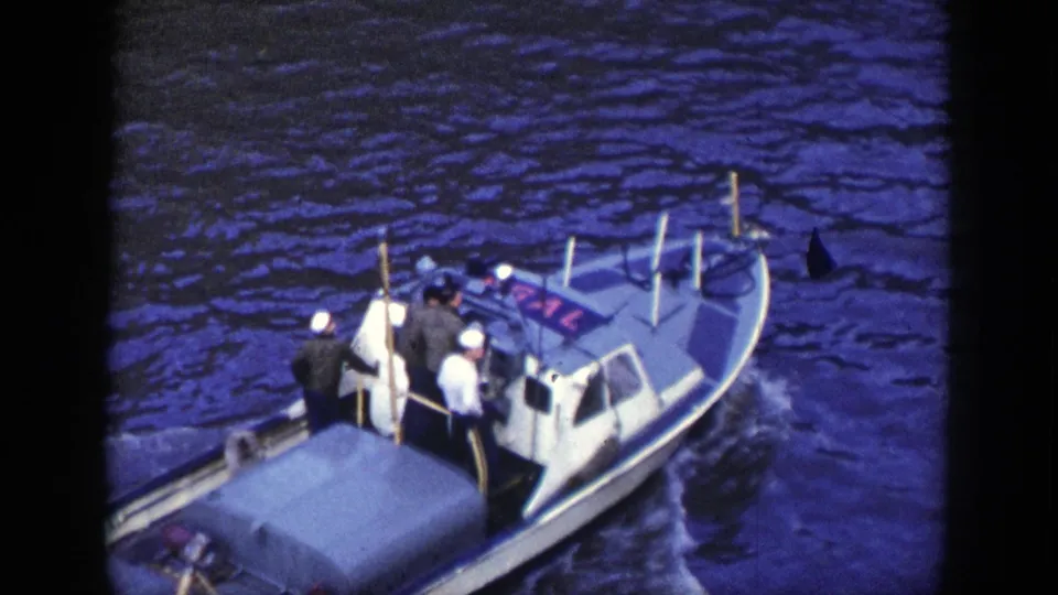 A small harbor motorboat or service craft maneuvers alongside a larger ship in the New York footage.