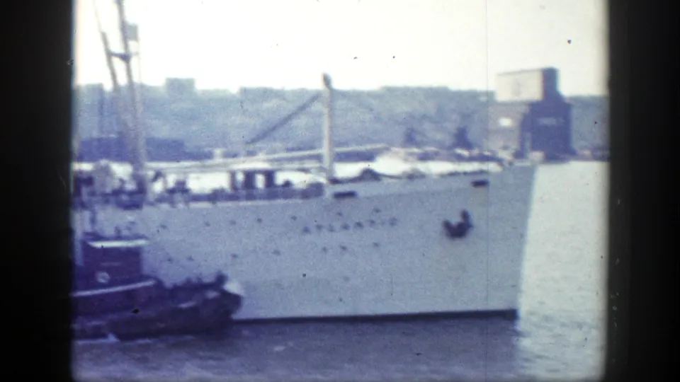 A large white ship hull with rows of portholes is seen in New York harbor, with a smaller tug or boat at the side.