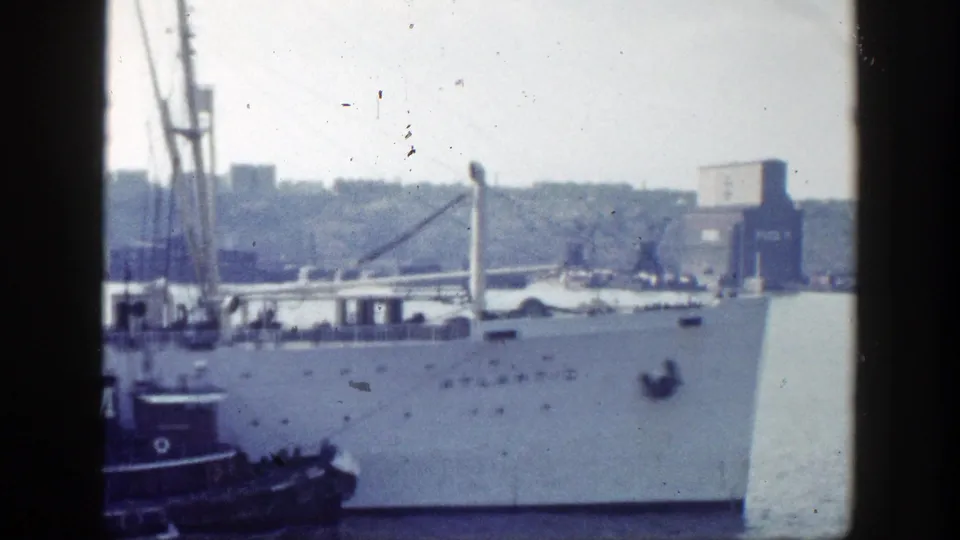 The large ship remains in frame with a small tug or harbor craft beside it and a hazy New York shoreline in the background.