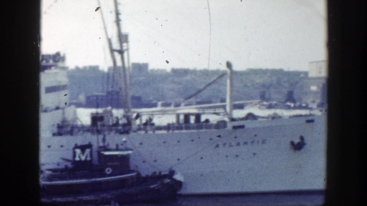 White passenger ship Atlantic beside a dark tugboat marked M in a harbor.