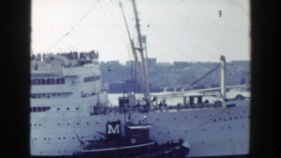 The Atlantic's superstructure and deck passengers appear as a tugboat works alongside in New York harbor.