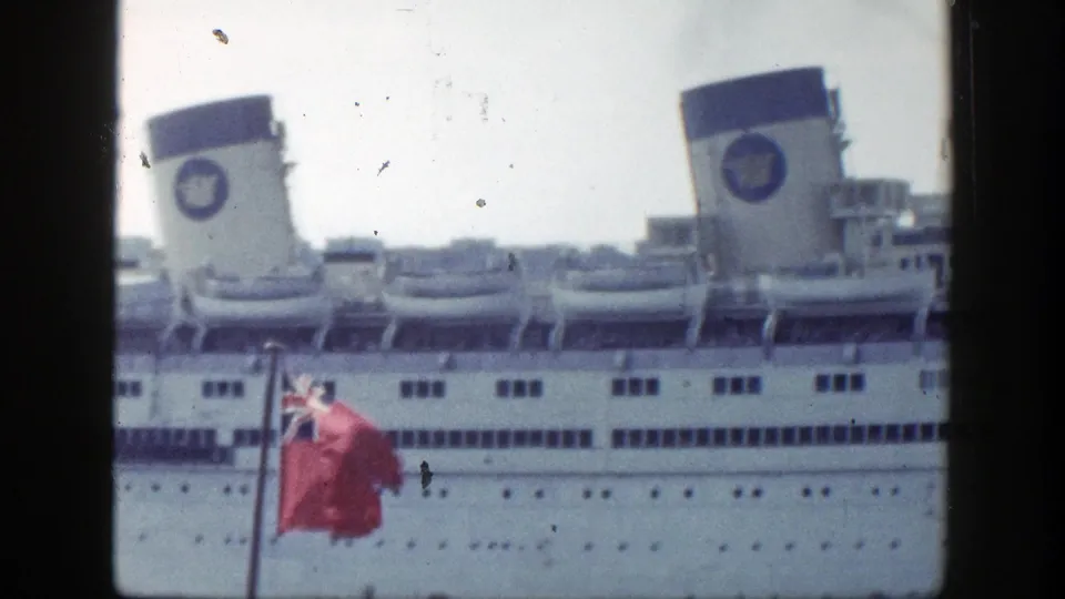 Soft 8mm frame from New York harbor showing the ocean liner's funnels, lifeboats, windows, and a red flag near the foreground edge.