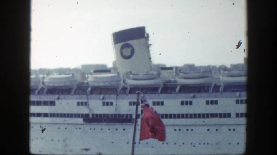 Color 8mm view of a docked white ocean liner in New York, with a red ensign flag partly visible in front.
