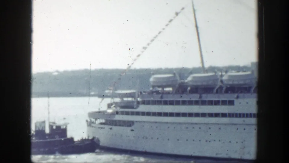 In New York harbor, a tugboat appears beside the hull of a large white ocean liner carrying rows of lifeboats on its upper deck.