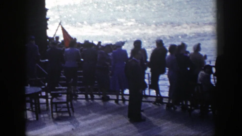 Passengers in 1948 New York harbor stand along a ship deck railing, watching the water near chairs and a red flag.
