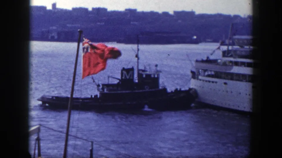 A tugboat sits beside a large passenger liner in New York harbor, with a red flag and distant skyline visible.