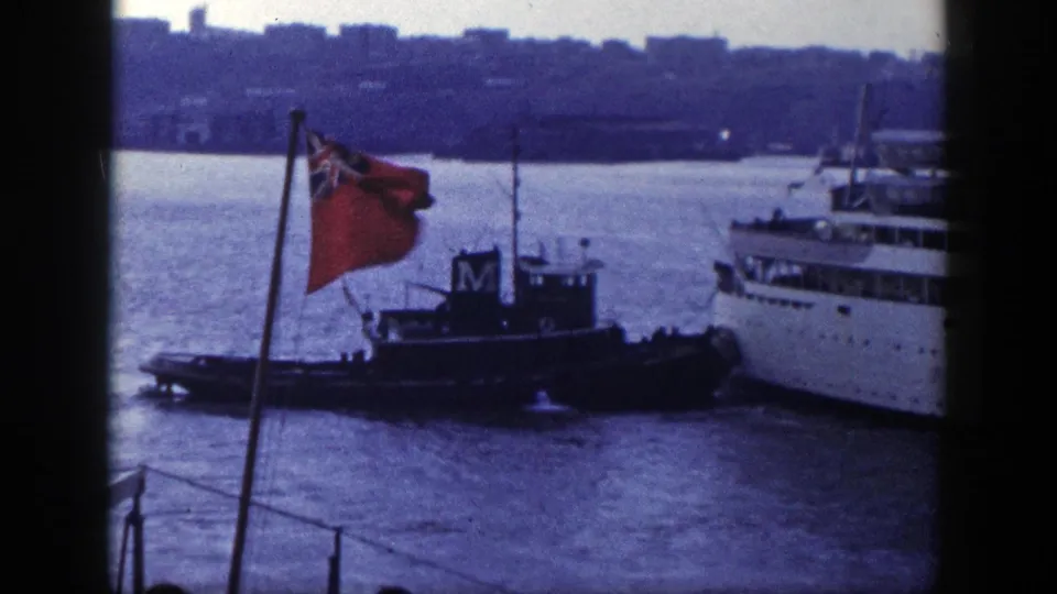 A steady deck view shows a tugboat beside a passenger liner in New York harbor, with a red flag moving in the foreground.
