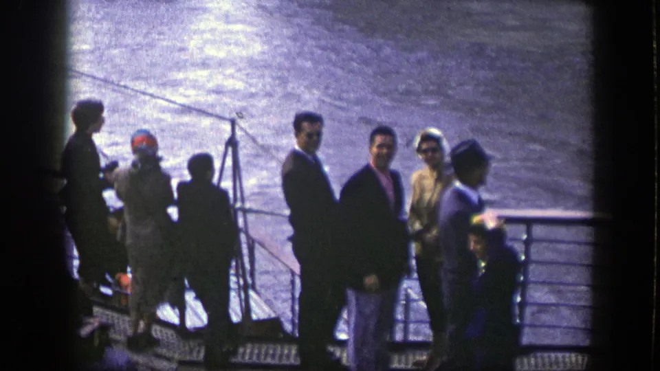 Passengers in suits, dresses, and hats stand by a ship railing with water beyond, likely during a New York cruise departure.
