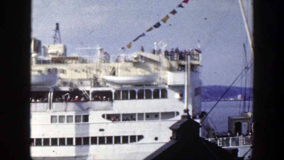 Large white passenger ship in New York harbor, with people gathered on upper decks and signal flags crossing the sky.