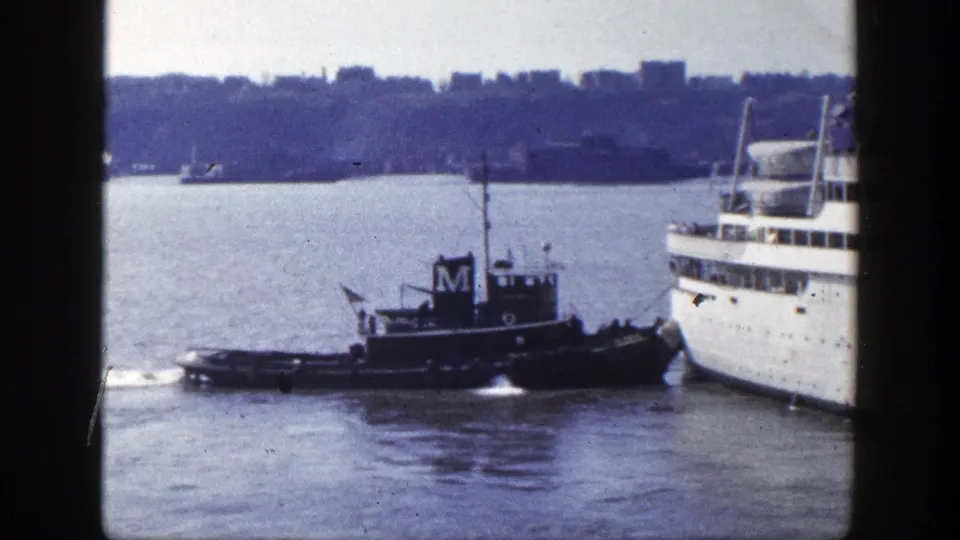 A dark tugboat marked with an M maneuvers alongside a large passenger ship in New York harbor, with shoreline visible across the water.