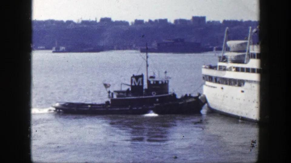 New York harbor scene showing a tugboat beside the passenger ship's bow, framed by dark vertical edges and a distant waterfront skyline.