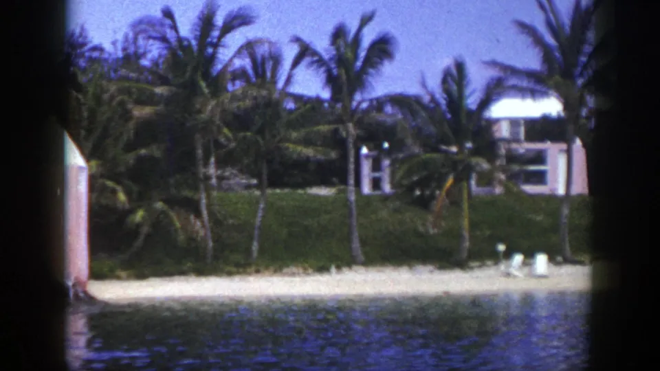 1948 8mm home-movie view of a palm-lined waterfront with blue water, sand, lounge chairs, and low buildings beyond a grassy bank.