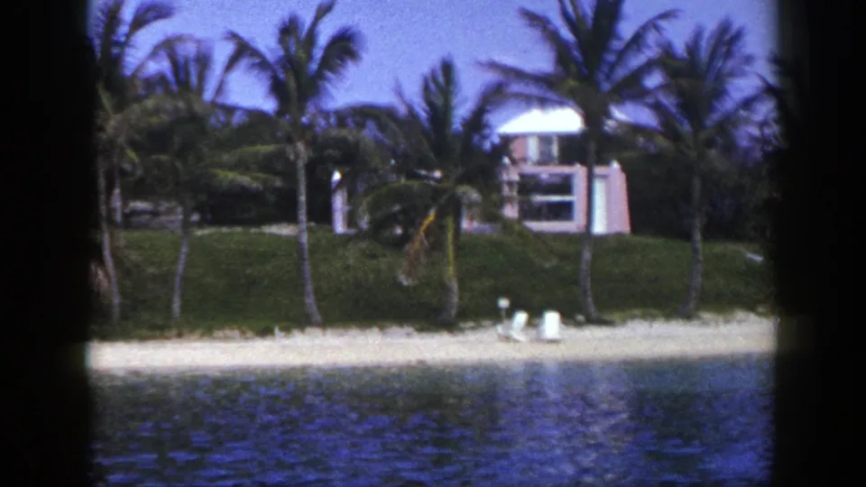 Palm trees frame a quiet 1948 waterfront scene with white lounge chairs on sand and pink-toned buildings above the beach.