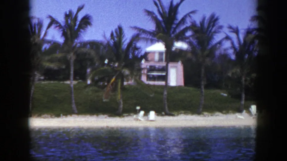 A soft 8mm frame shows palm trees, a grassy bank, beach chairs, and a low waterfront building in the 1948 home-movie scene.
