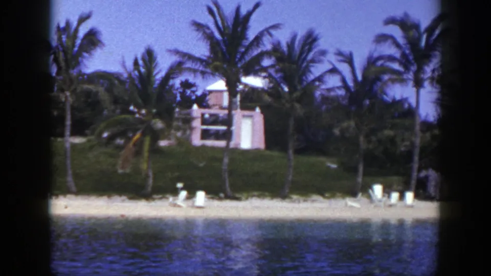 1948 8mm waterfront view with palm trees, white beach chairs, rippling water, and a pink-toned building on the grassy rise.