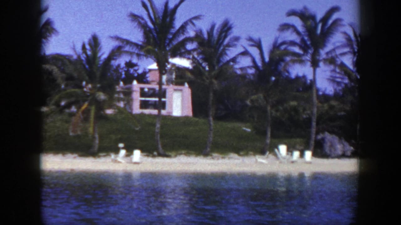 Beachfront with palms, lounge chairs, water, and a pink building on a grassy rise.