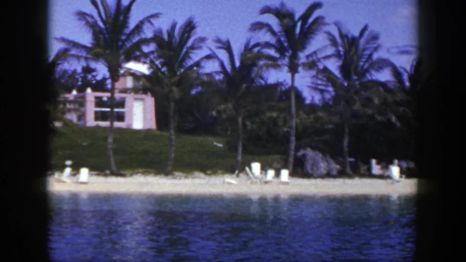 1948 home-movie frame of a tropical-looking shoreline with palms, lounge chairs, rocks, water, and a pink building behind the beach.