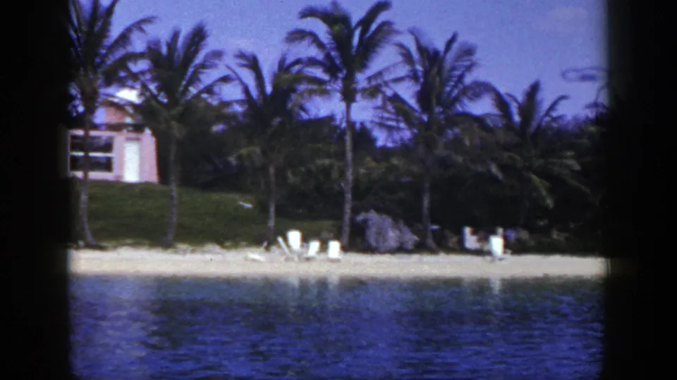 1948 8mm frame during the pan, showing blue water, a sandy beach, palms, lounge chairs, rocks, and part of a pink building.