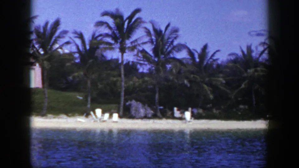 1948 home-movie frame showing the right side of the beach pan, with palms, lounge chairs, rocks, sand, and calm water.