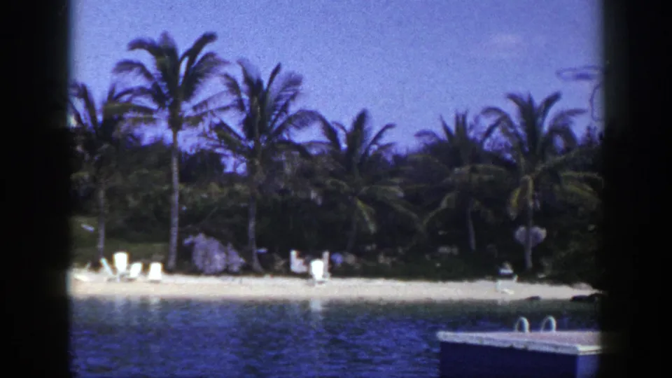 1948 8mm frame with a tropical-looking beach, dense palms, white lounge chairs, and part of a floating dock in the foreground.